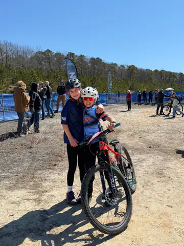Two smiling riders hug beside a mountain bike at a race on a sunny day.