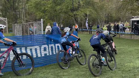 Shredders riders pedal around a grassy corner during a race, with families watching from the fence.
